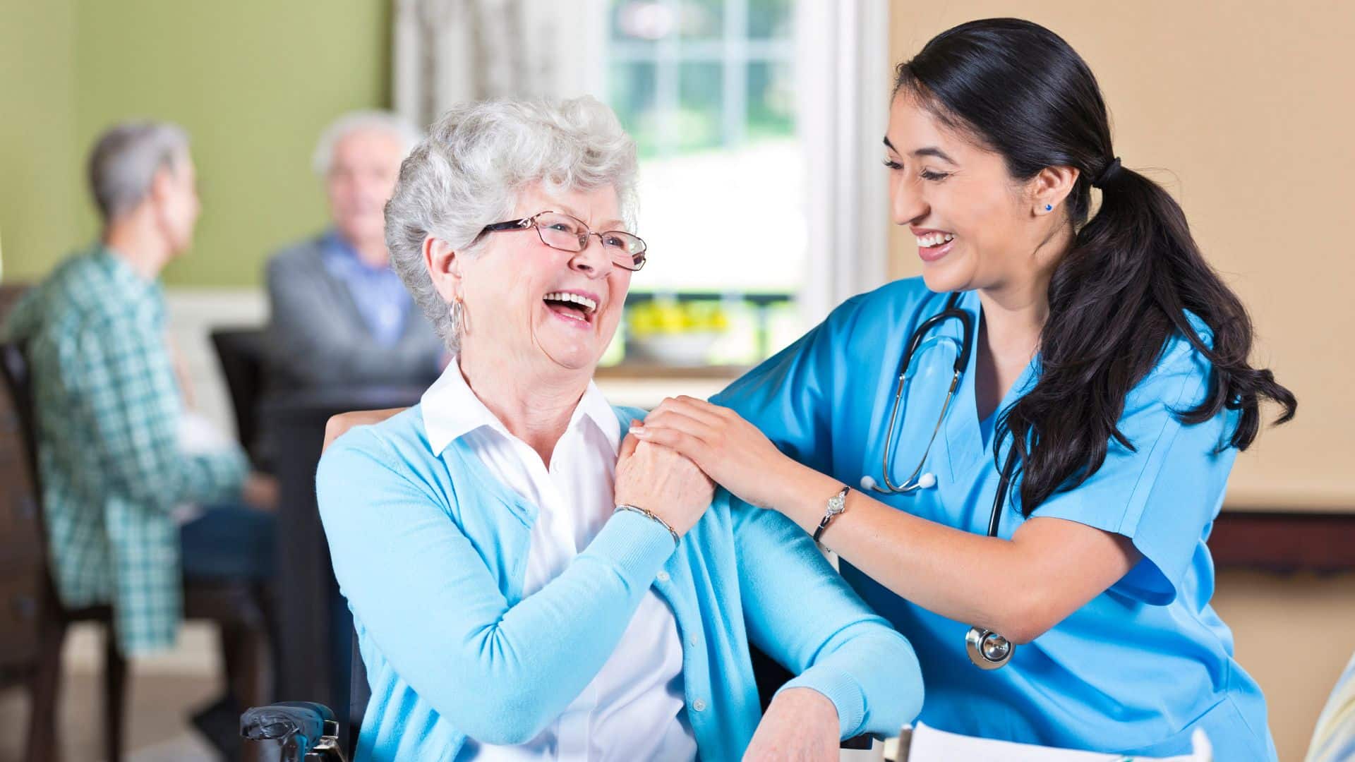 female care professional holding hand of elderly women sitting in a chair both laughing