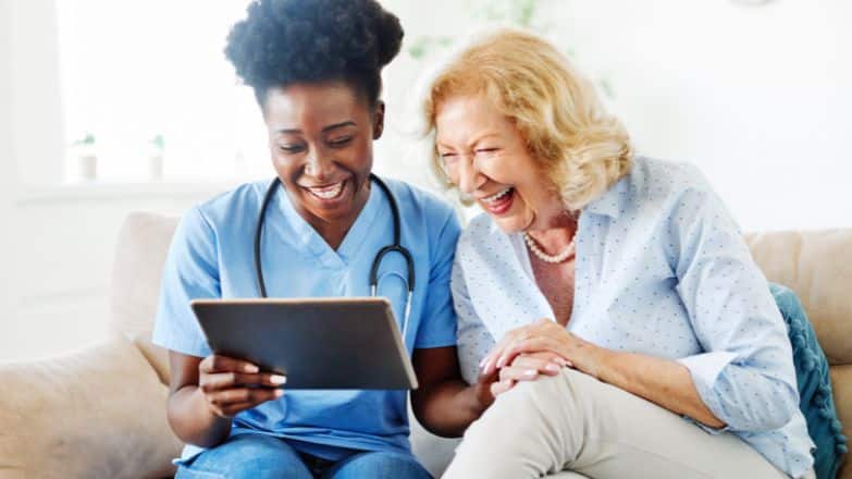 Nurse and elderly women sitting on couch looking at a tablet laughing